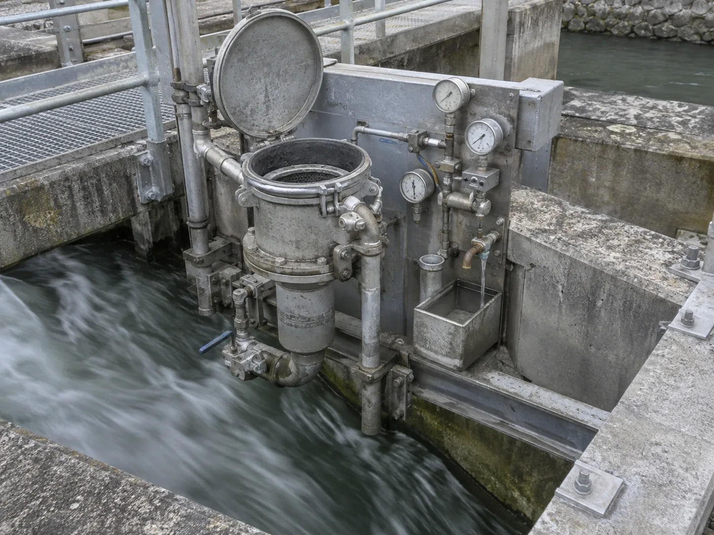 Close photograph of an industrial water sampling station on a concrete channel: a steel inspection hatch open on a flowing conduit, with a filter housing, gauges and a small sample tap mounted beside it. The equipment is utilitarian, weathered, institutional gray and galvanized steel.