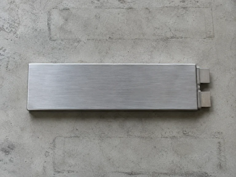 Clean overhead photograph of a single flat rectangular battery cell lying on a pale concrete workbench: a long slim prismatic cell in brushed aluminum with two plain terminals at one end. Nothing else on the bench except the faint outlines where other cells have sat.