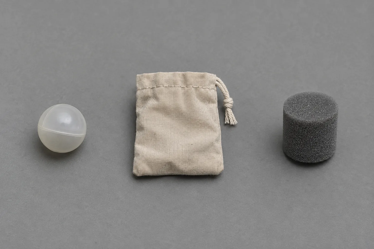 Overhead studio photograph on a neutral mid gray seamless background: three plain objects laid out in a row with generous even spacing, like a museum specimen tray. Left, a small hollow plastic sphere about the size of a marble.