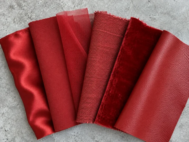 Close overhead photograph of six fabric swatches fanned out on a pale gray workbench, all of them the same saturated warm red but in six different materials: silk satin, matte wool crepe, fine tulle, coarse linen, velvet and smooth leather. Each takes the dye and the light differently, so the same red reads as six shades.