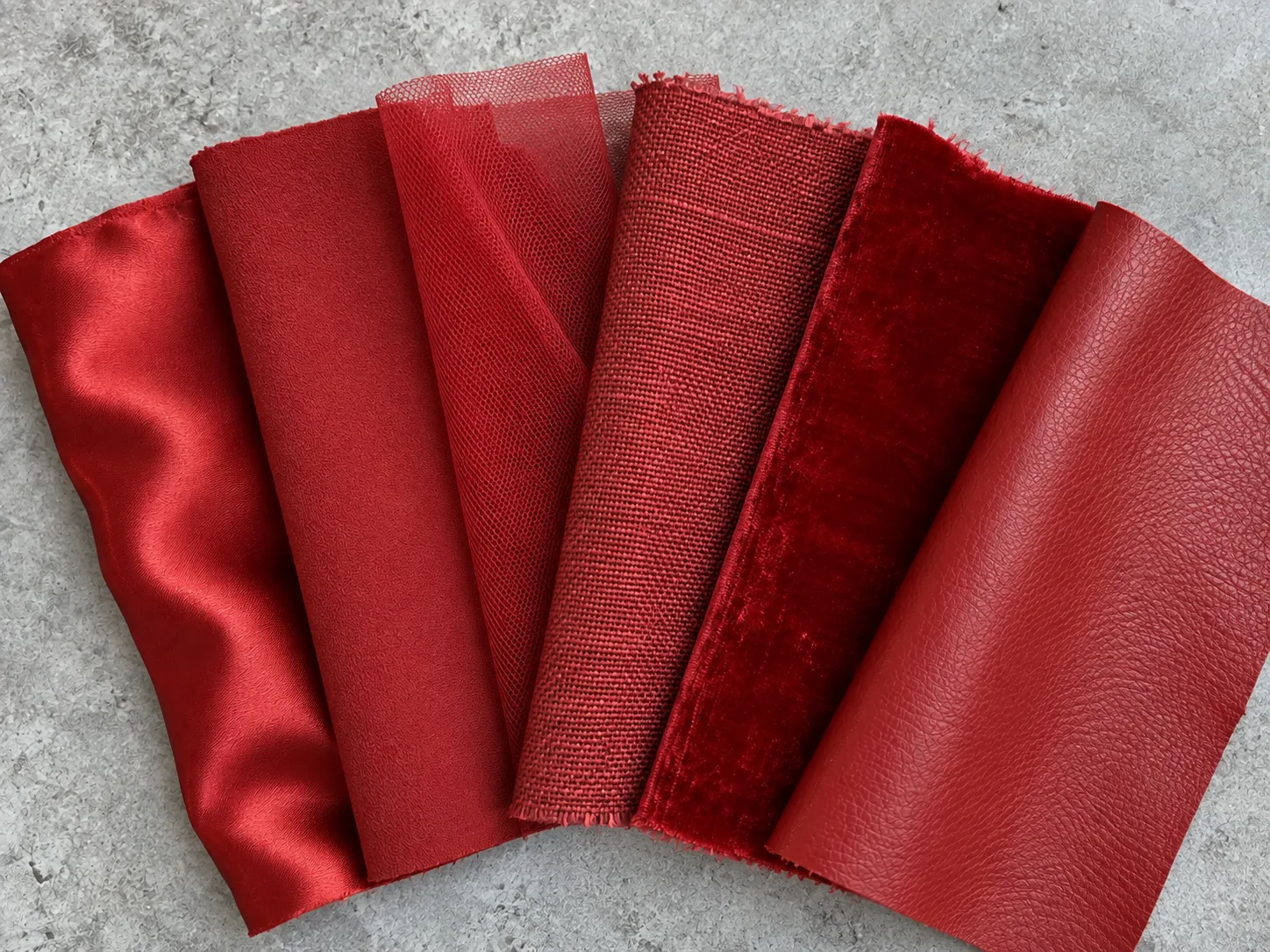 Close overhead photograph of six fabric swatches fanned out on a pale gray workbench, all of them the same saturated warm red but in six different materials: silk satin, matte wool crepe, fine tulle, coarse linen, velvet and smooth leather. Each takes the dye and the light differently, so the same red reads as six shades.