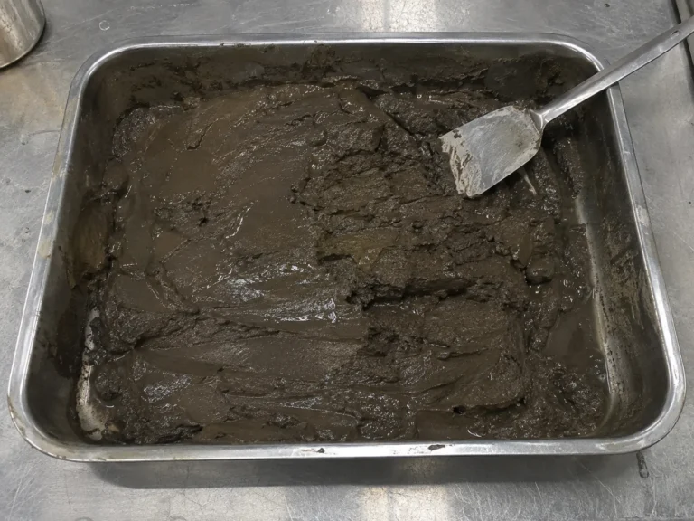 Close overhead photograph of a shallow stainless steel tray on a scuffed lab bench, holding a few kilograms of wet deep sea sediment: fine grained, dark brown gray, smooth and slightly glossy with seawater, with a clean spatula laid across one corner. The tray is plain and unlabelled.