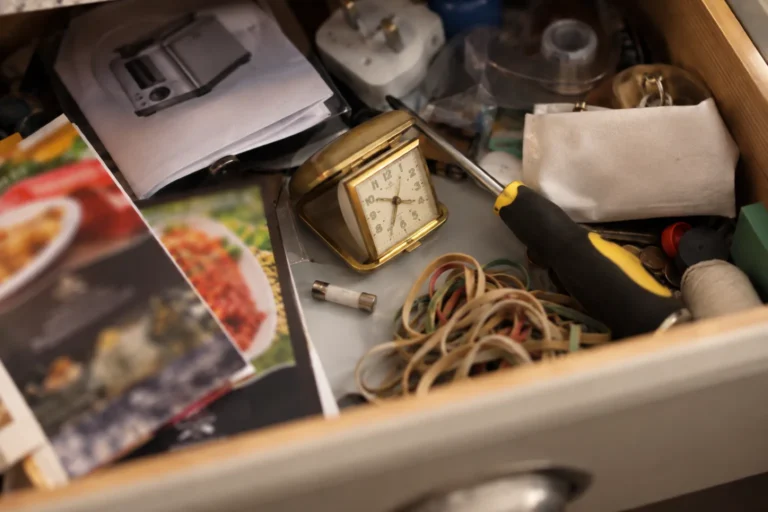Photograph looking down into an open kitchen drawer, the domestic one that holds everything: a folded appliance booklet, a spare fuse, a tangle of elastic bands, two takeaway menus with the print too soft to read, a screwdriver, and a small brass travel alarm clock lying on its side among them.