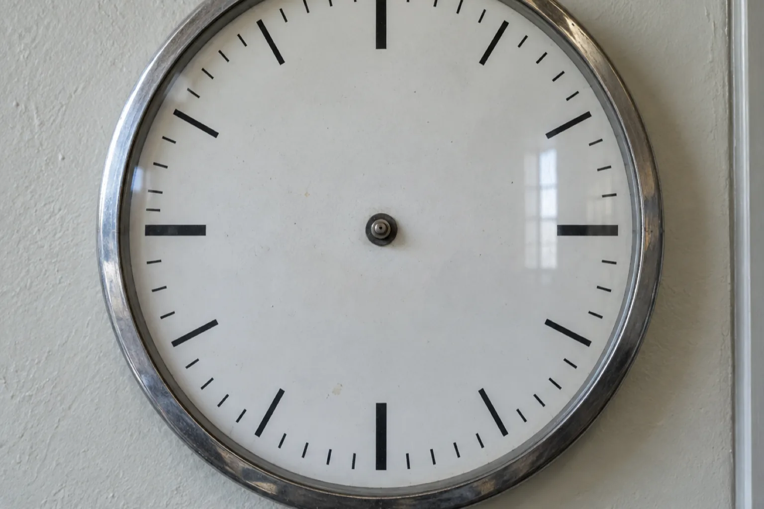 Close photograph of a large institutional wall clock with both hands removed, leaving only the bare center spindle on an otherwise ordinary white face. The markings are plain, the glass slightly dusty, the chrome bezel dulled.