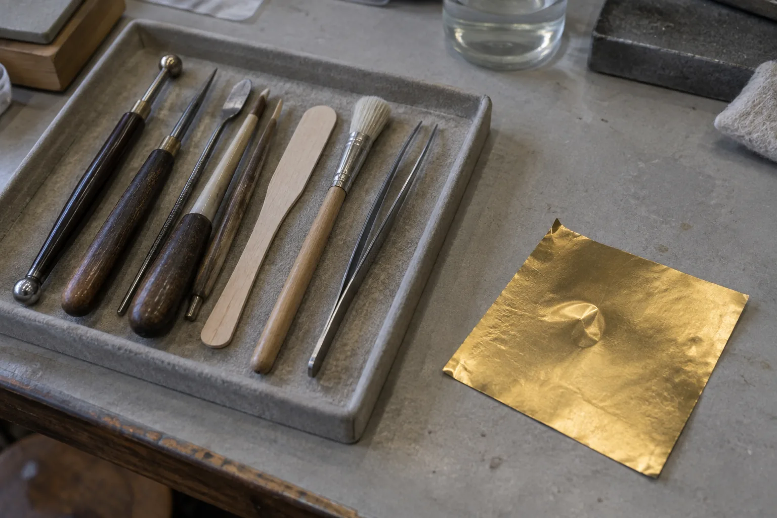 Close photograph of a conservator's bench: a shallow tray holding small hand tools laid in a row, burnishers, a wooden spatula, a soft brush, a pair of fine tweezers, all worn with use.