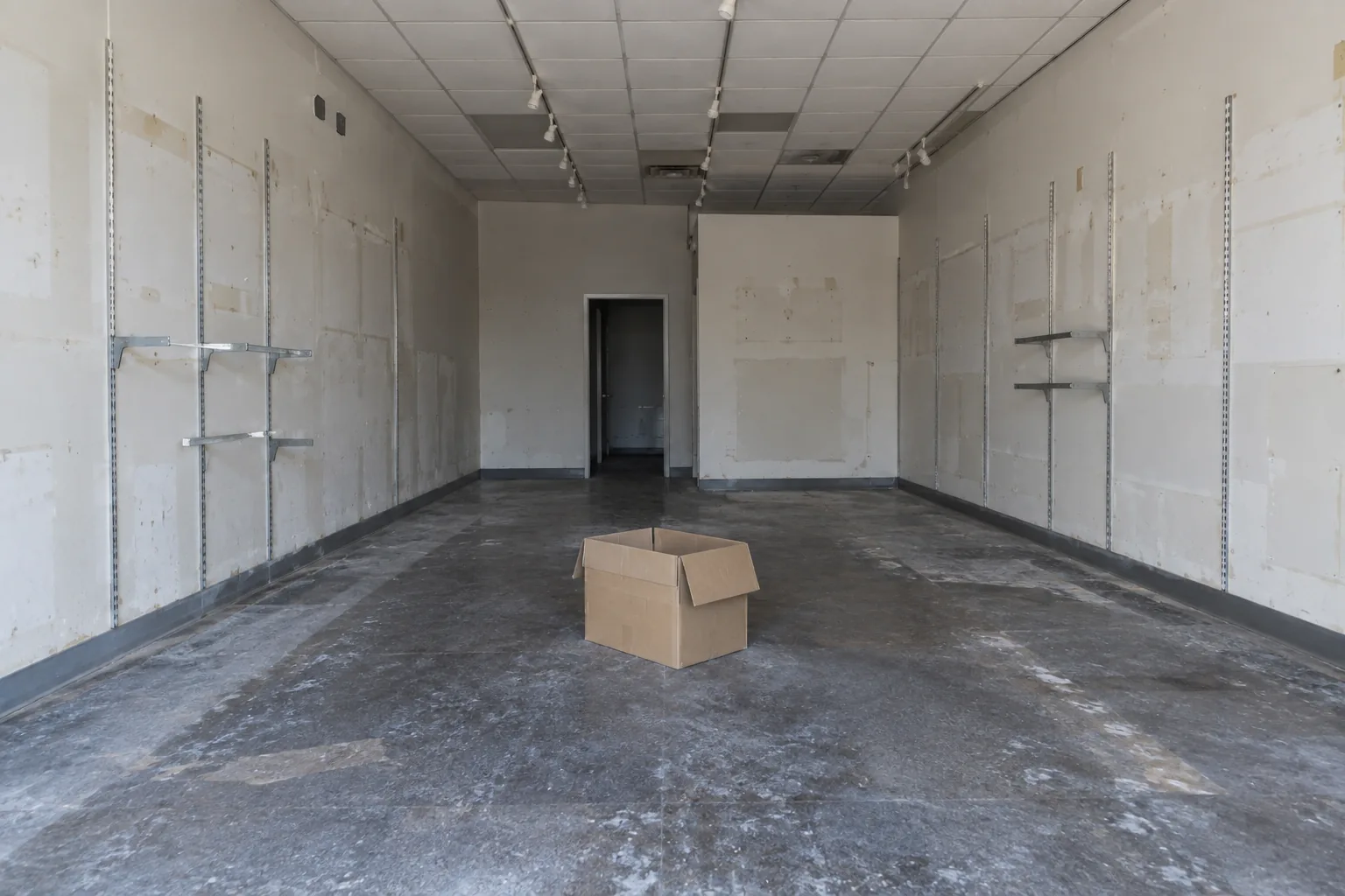Photograph of an empty retail shop unit, cleared out. Bare walls with pale rectangles and screw holes where signage and shelving used to be, exposed brackets still fixed in places, a scuffed floor, one empty cardboard box left in the middle.