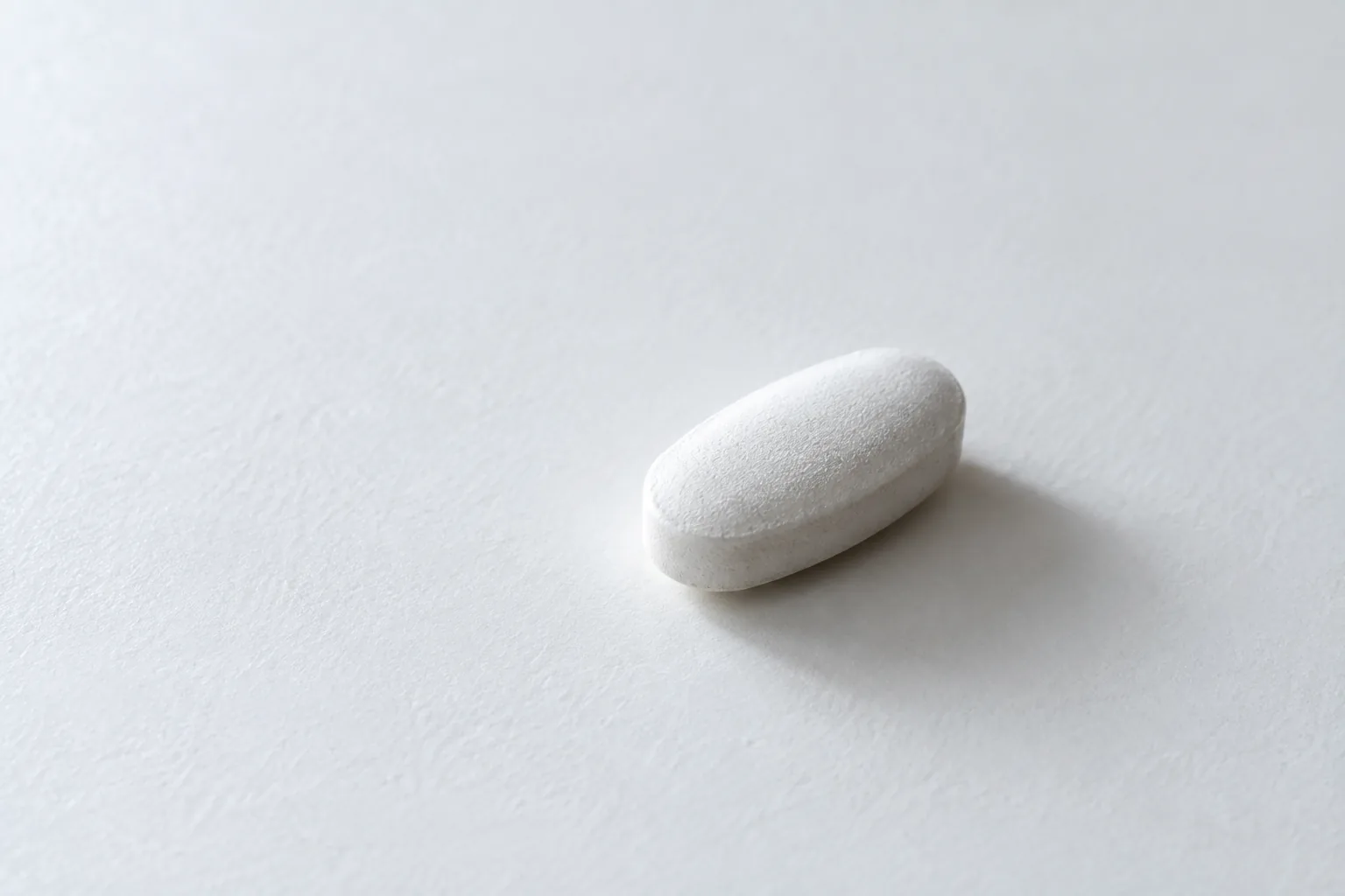 Extreme close macro photograph of a single plain white oval tablet resting on a large expanse of matte white surface, with nothing else in frame. The tablet is unmarked, no score line, no imprint.