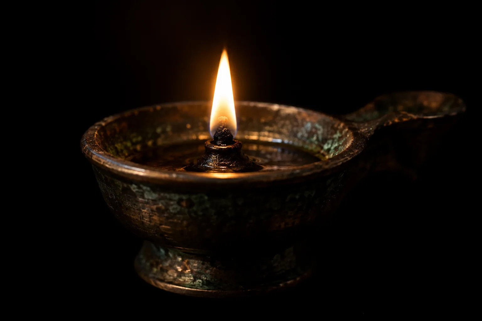 Photograph of a small aged bronze oil lamp with a side handle, burning with one steady upright flame against a completely black background. The metal is pitted and shows green patina.