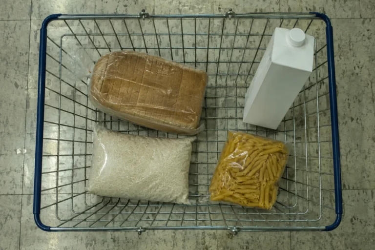 Overhead photograph of a wire supermarket basket on a concrete floor holding four plain items with no branding: a wrapped sliced loaf, a white carton, a clear bag of rice and a bag of penne pasta.