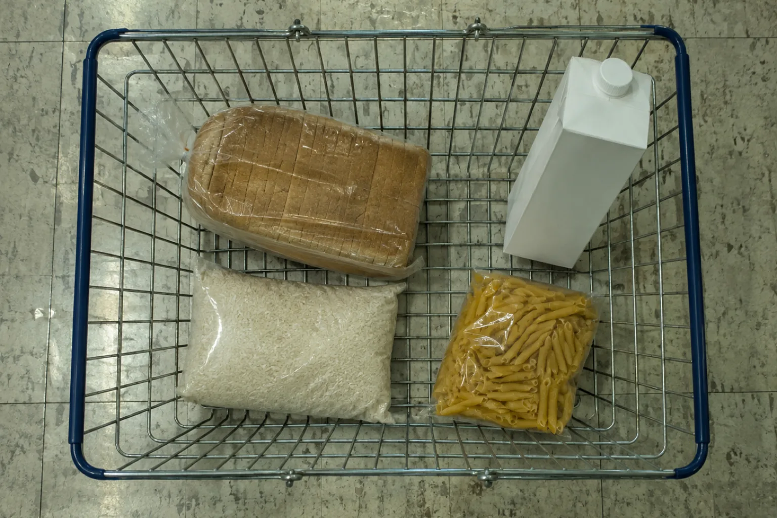 Overhead photograph of a wire supermarket basket on a concrete floor holding four plain items with no branding: a wrapped sliced loaf, a white carton, a clear bag of rice and a bag of penne pasta.