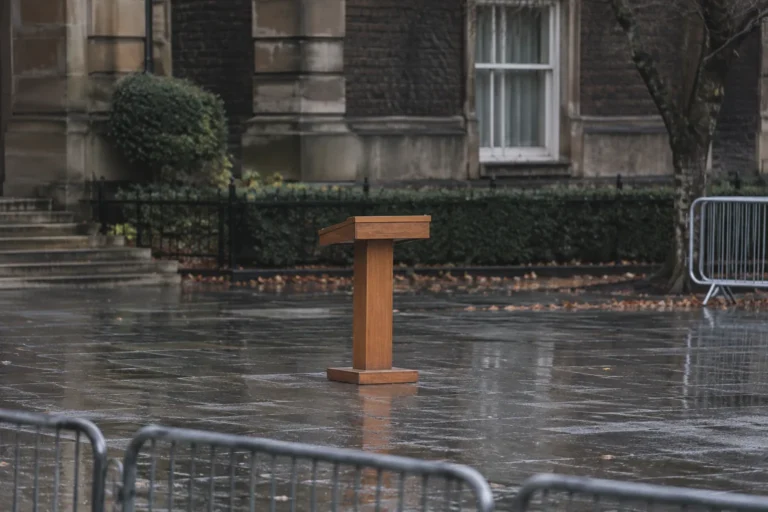 Photograph of a plain wooden lectern standing alone on wet pavement outside a stone government building on a gray day, with metal crowd barriers in the foreground and nobody anywhere in the frame.