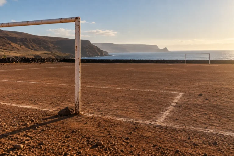 Photograph of a bare dirt football pitch beside the sea, its lines marked in white powder and its two goals standing empty, the nearer frame rusted through its white paint. Low cliffs and evening light fill the background.