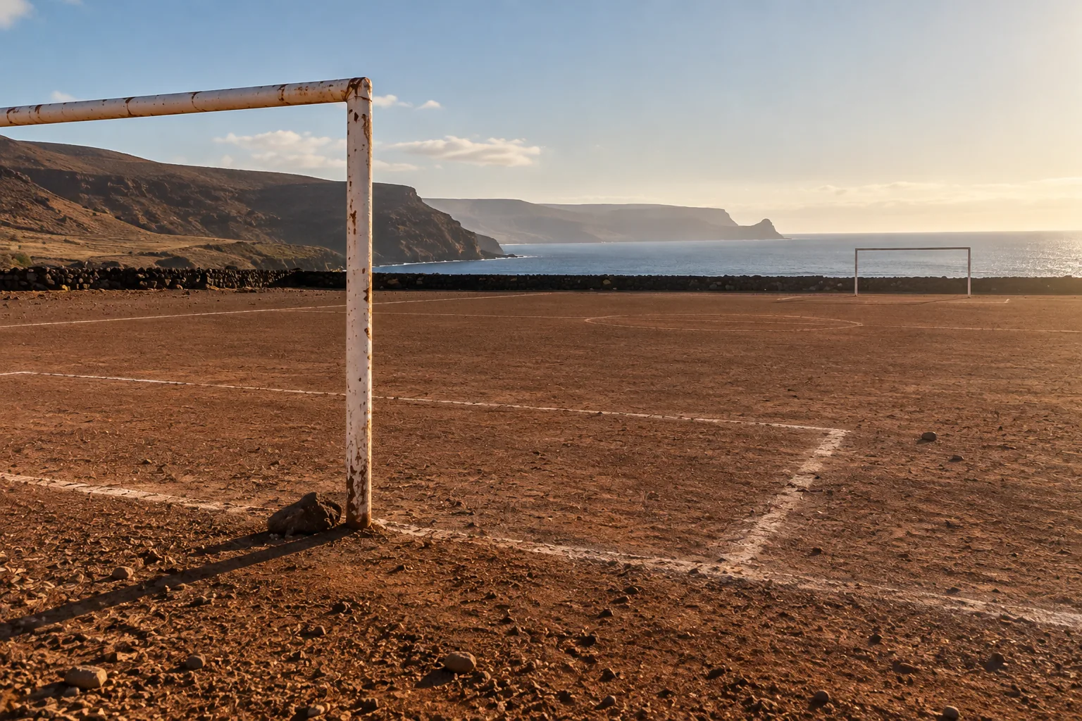 Photograph of a bare dirt football pitch beside the sea, its lines marked in white powder and its two goals standing empty, the nearer frame rusted through its white paint. Low cliffs and evening light fill the background.
