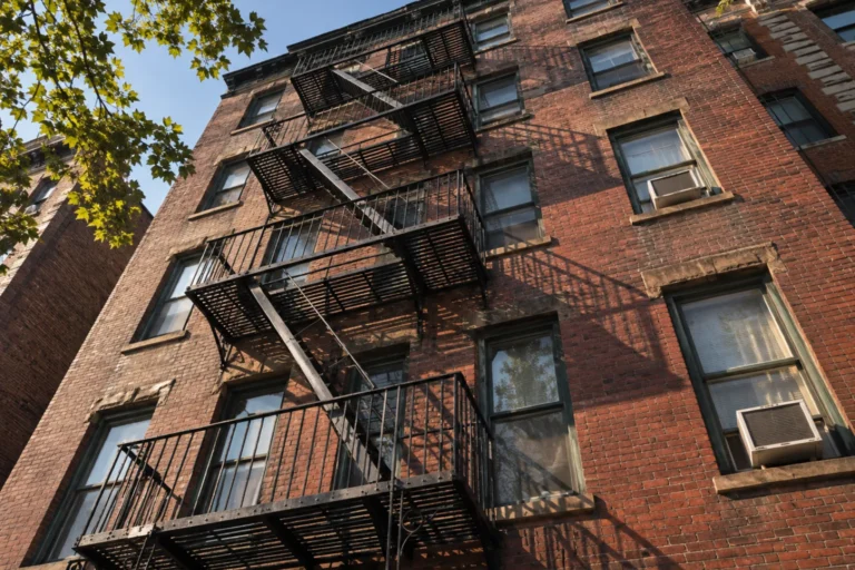 Photograph looking up the face of an older brick apartment building, its black iron fire escapes zigzagging across the windows and window mounted air conditioning units set in several of the openings.