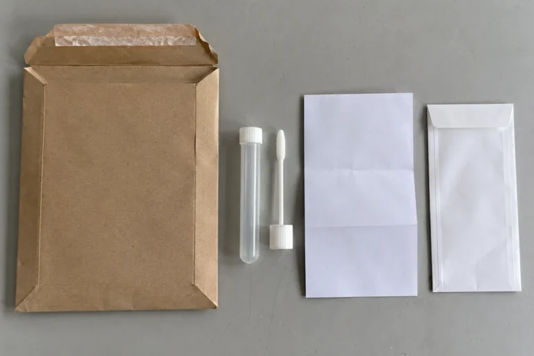 Overhead photograph of the contents of a mail in sample kit laid out on a gray surface: a padded brown envelope, a sealed plastic collection tube, a swab, a folded blank instruction sheet and a small white return envelope.