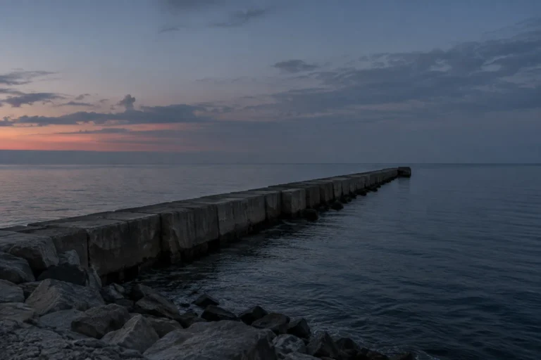 Photograph of a long concrete breakwater running out from a rocky shore into calm open water at dusk, seen from the land side. The weathered blocks recede toward a hazy horizon under dim blue evening light, and there are no people or vessels anywhere.