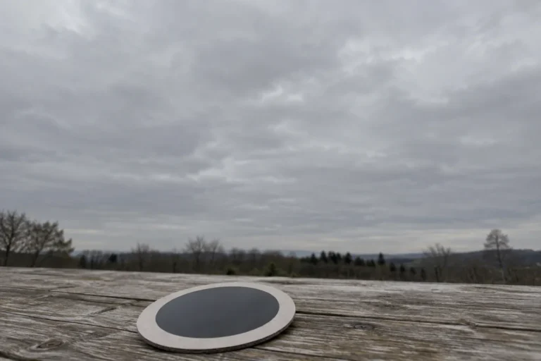 Photograph of a small circular solar viewing filter in a plain cardboard mount lying alone on a weathered wooden table outdoors. Above and behind it a large expanse of flat gray overcast sky fills most of the frame, with no sun visible at all.