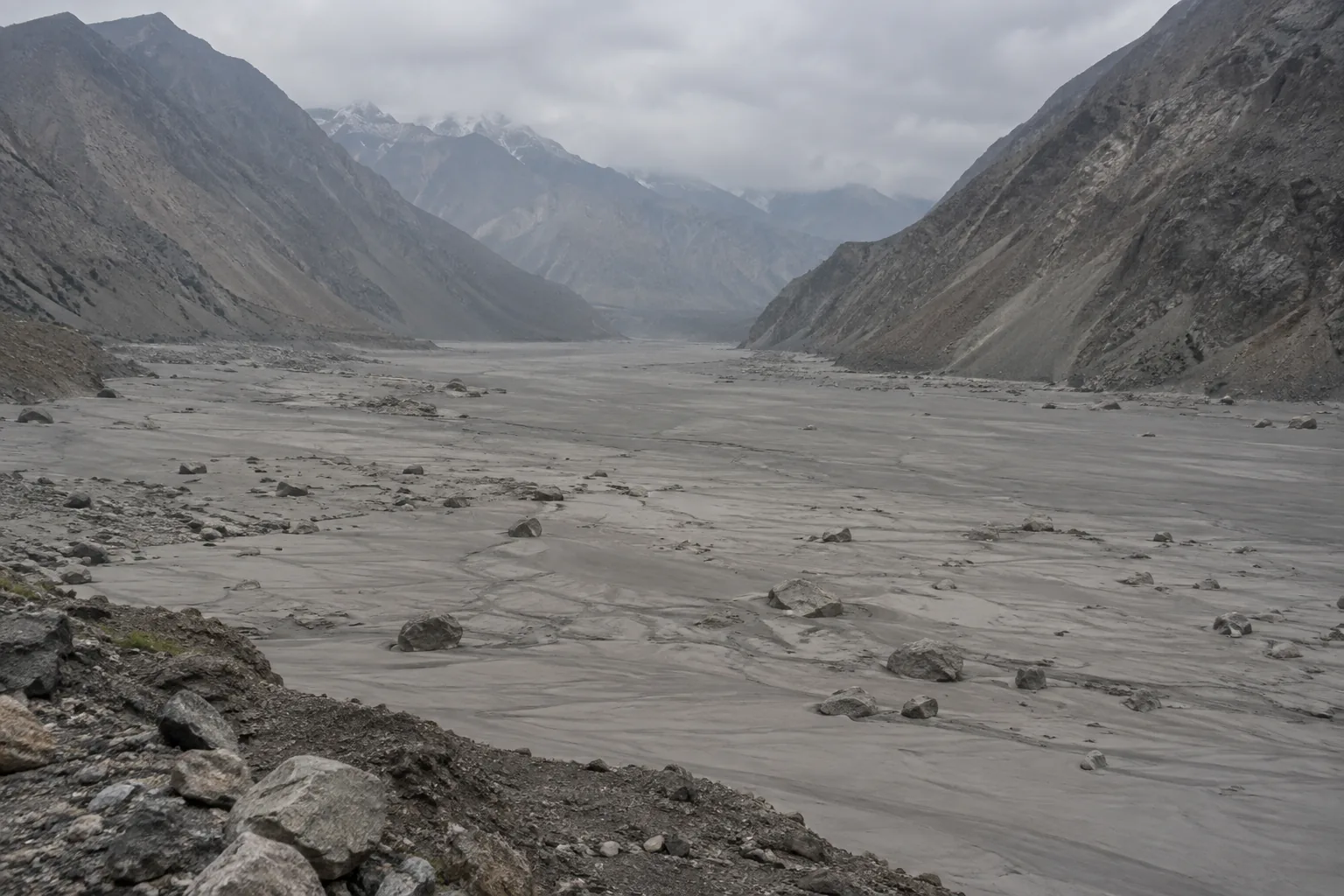 Wide photograph of a broad river valley floor covered in fresh gray sediment and scattered boulders, the surface smooth and featureless where the deposit has settled. Steep valley walls rise on both sides under flat overcast light, with no water and no vegetation on the deposit.