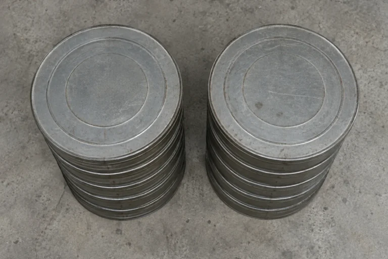 Overhead photograph of two identical stacks of plain unlabeled film reel cans in flat gray metal standing side by side on a concrete floor. Each stack is about six cans high and exactly the same height as the other, the cans scuffed and completely blank.