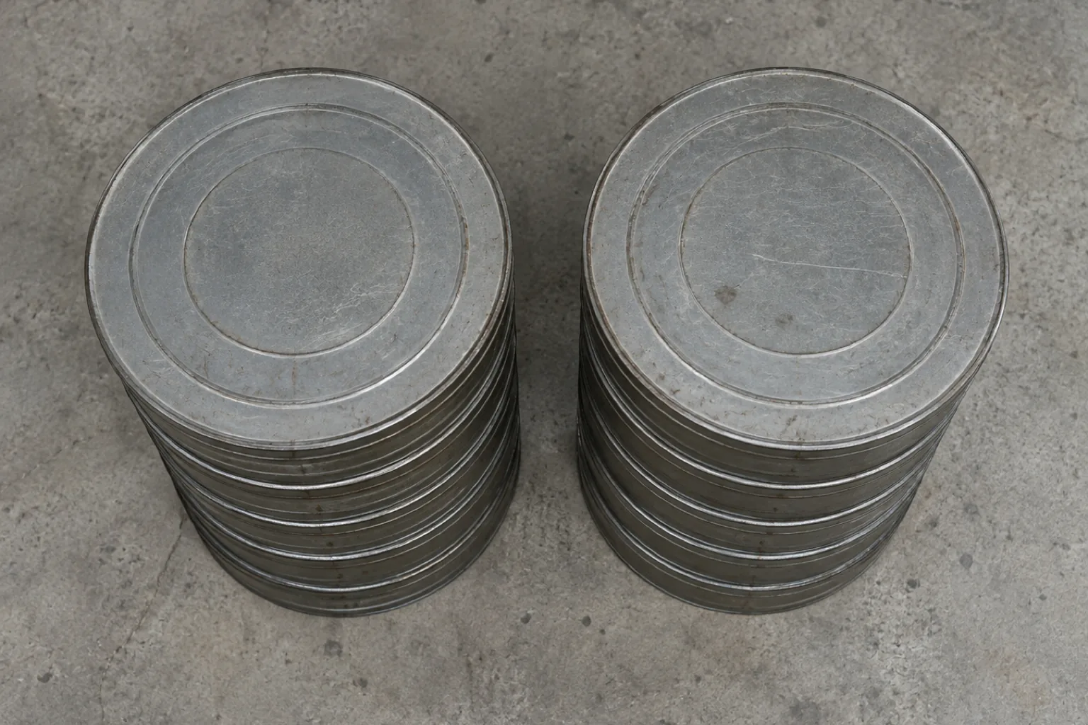 Overhead photograph of two identical stacks of plain unlabeled film reel cans in flat gray metal standing side by side on a concrete floor. Each stack is about six cans high and exactly the same height as the other, the cans scuffed and completely blank.