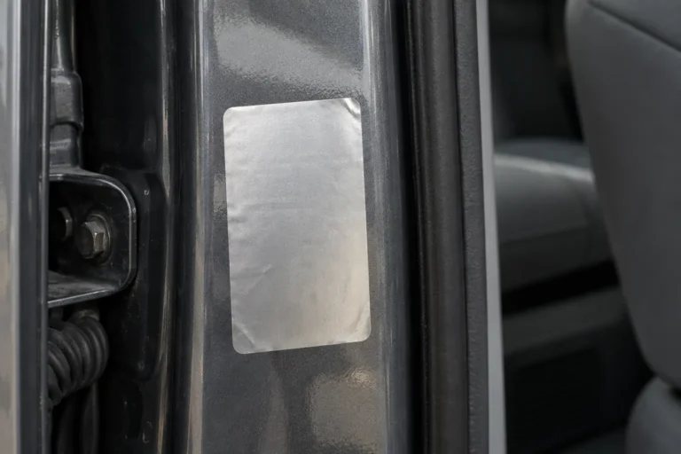 Tight close photograph of a blank silver foil compliance label stuck to the painted metal edge of an open car door frame, slightly creased at one corner. There is no printing on it of any kind, and a sliver of plain gray interior sits out of focus beyond.
