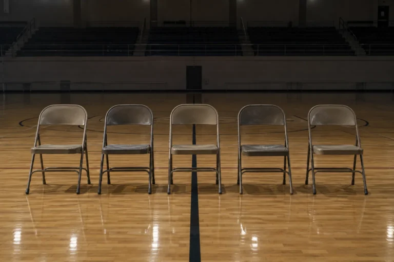 Photograph of five empty folding chairs in a row on a polished wooden floor in an otherwise empty sports hall, shot straight on. The house lights are low, the seating tiers behind fall into shadow, and the court markings run under the chairs.